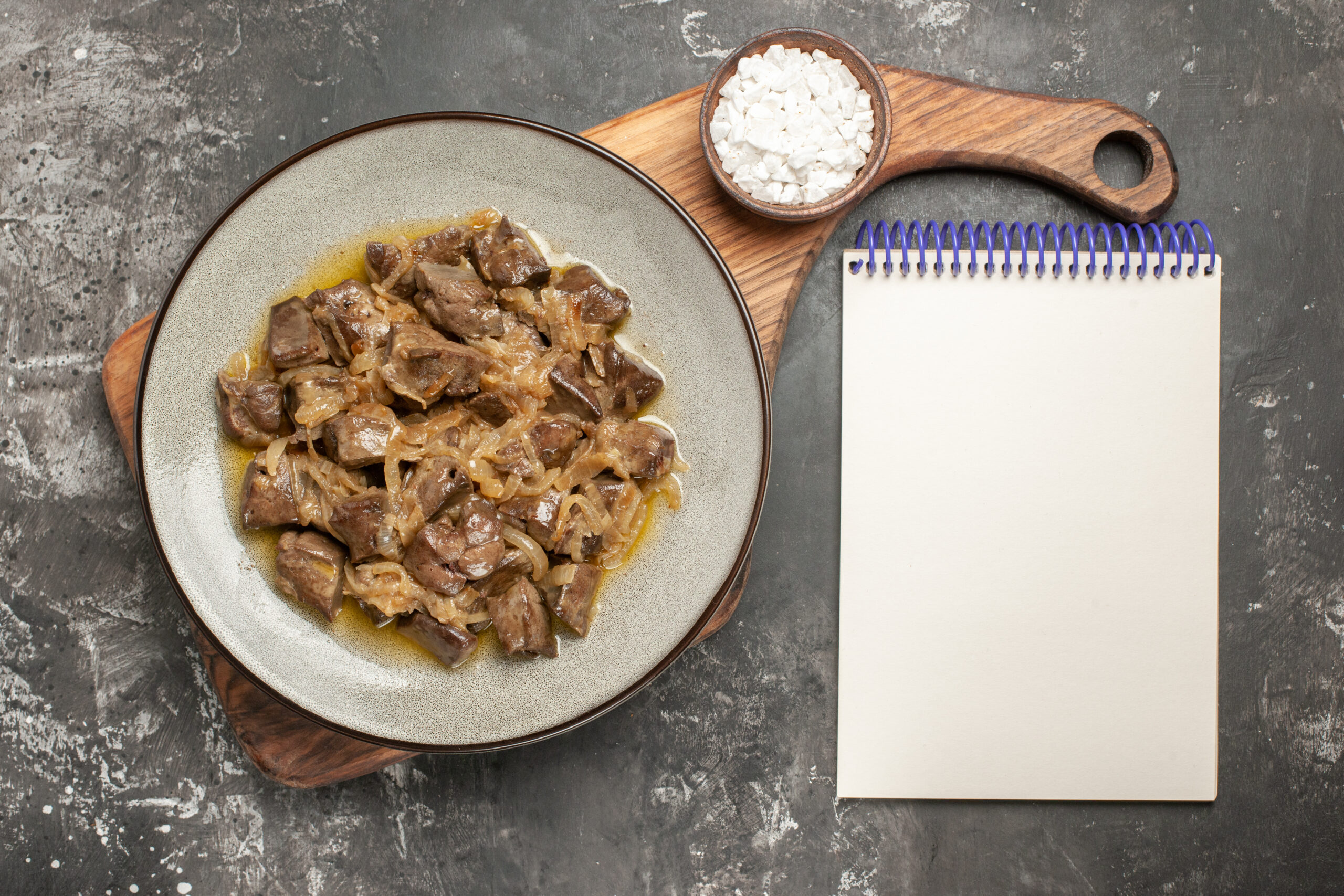 A plate of sautéed chicken livers with caramelized onions, placed on a wooden board with a bowl of sea salt and a blank notebook on a dark textured background.