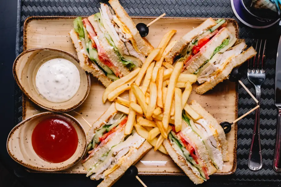 Four triangular slices of club sandwich with fresh vegetables, deli meat, and cheese, served with French fries in the center and bowls of ketchup and mayonnaise on the side.