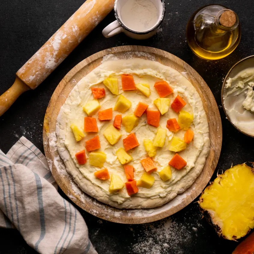 A raw fruit pizza crust topped with cubed pineapple and papaya, placed on a wooden board with baking ingredients like flour, oil, and a rolling pin.