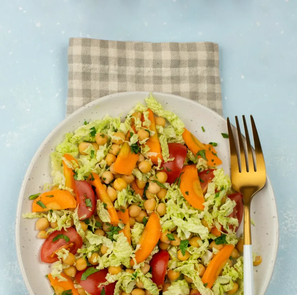 A colorful chickpea salad with napa cabbage, cherry tomatoes, sliced carrots, and fresh parsley served in a white bowl with a gold-handled fork.