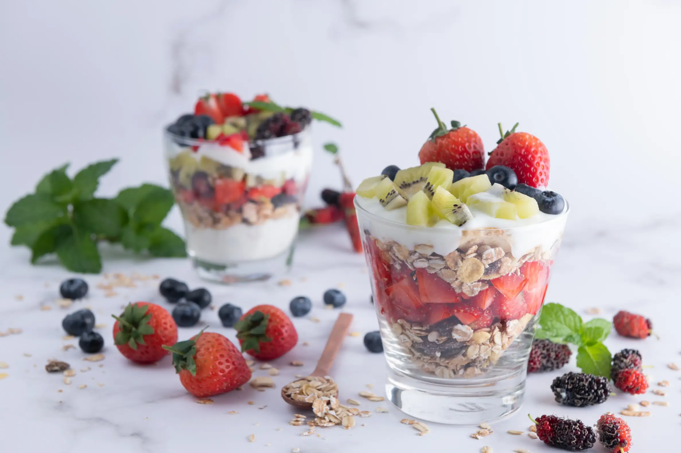 Two glasses of layered yogurt parfaits with Greek yogurt, strawberries, blueberries, kiwi, granola, and oats, garnished with whole strawberries and mint leaves, placed on a white marble surface with scattered berries and oats.