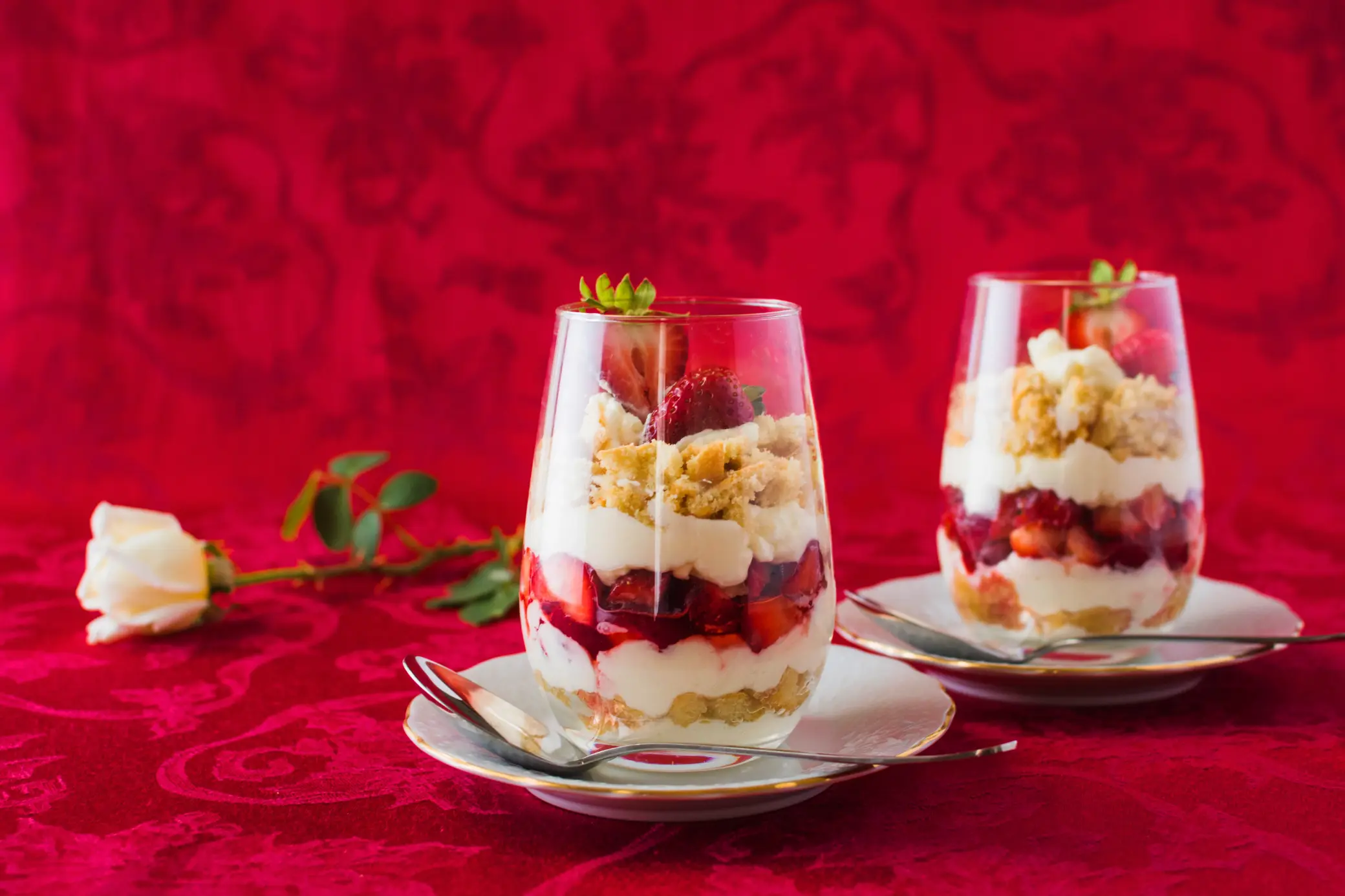 Two elegant glasses of layered yogurt parfait with Greek yogurt, fresh strawberries, crumbled granola, and cake pieces, served on white plates with spoons, against a red fabric background with a white rose beside them.