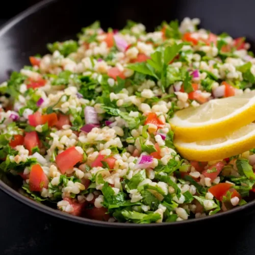 A bowl of quinoa tabbouleh with chopped parsley, tomatoes, red onions, and lemon slices, garnished with fresh herbs.