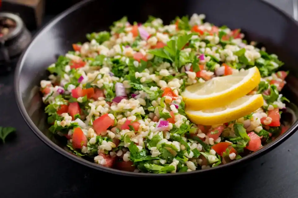 A bowl of quinoa tabbouleh with chopped parsley, tomatoes, red onions, and lemon slices, garnished with fresh herbs.