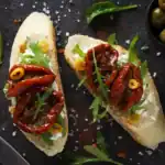 Toasted baguette slices topped with creamy cheese, sun-dried tomatoes, arugula, and sliced olives on a dark rustic surface.
