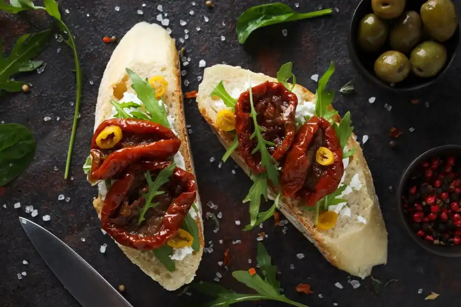 Toasted baguette slices topped with creamy cheese, sun-dried tomatoes, arugula, and sliced olives on a dark rustic surface.
