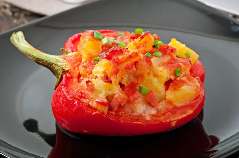 A halved red bell pepper stuffed with a cheesy mixture of potatoes, tomatoes, and green onions, baked to a golden brown and served on a black plate.