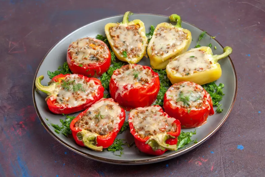 Red and yellow bell peppers stuffed with meat and rice, topped with melted cheese and fresh herbs, served on a gray plate.