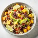 A vibrant quinoa salad with black beans, mango, avocado, cherry tomatoes, and lime slices in a white bowl.