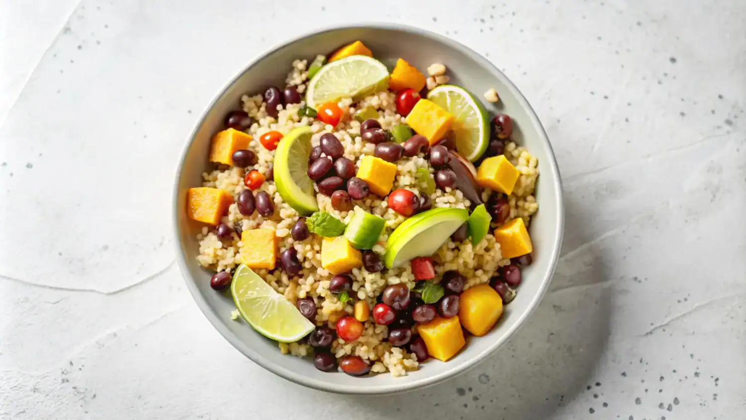 A vibrant quinoa salad with black beans, mango, avocado, cherry tomatoes, and lime slices in a white bowl.