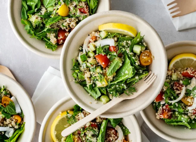 Bowls of fresh quinoa salad with spinach, cherry tomatoes, cucumber, herbs, and lemon slices, served with wooden forks.