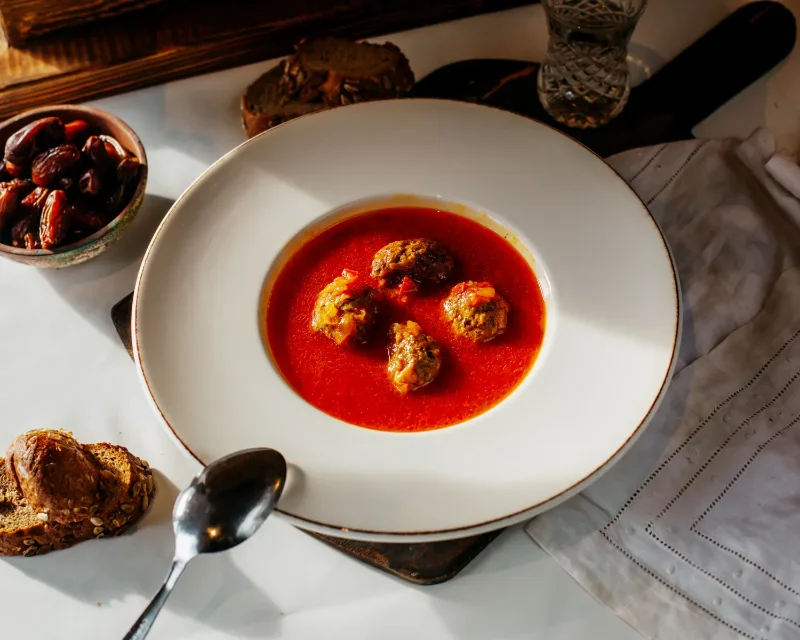 A white bowl filled with traditional Italian meatballs served in a rich tomato sauce, placed on a rustic table with sliced bread, dates, and a glass of water.