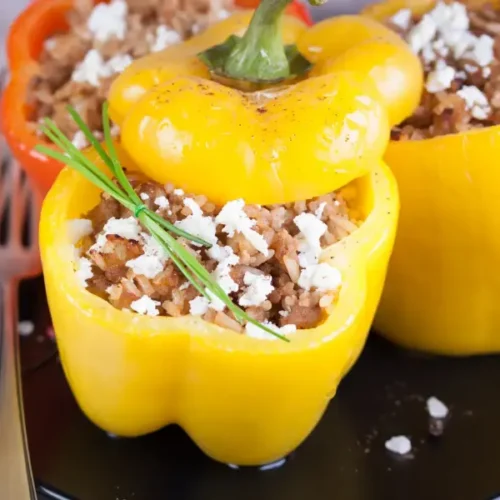 Colorful bell peppers stuffed with ground beef, rice, and crumbled cheese, garnished with fresh herbs on a black plate.