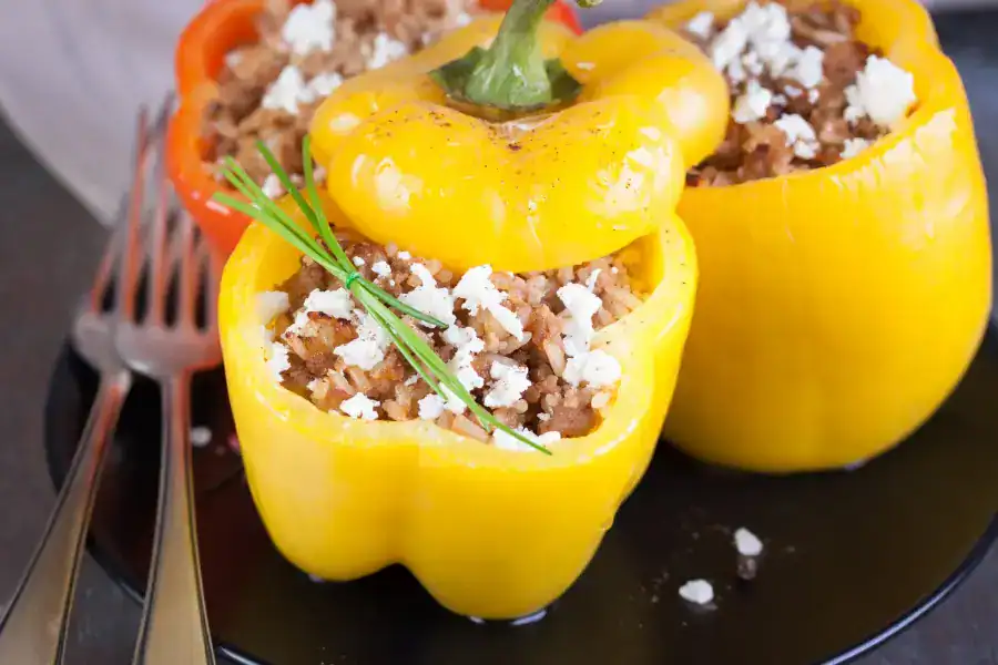 Colorful bell peppers stuffed with ground beef, rice, and crumbled cheese, garnished with fresh herbs on a black plate.