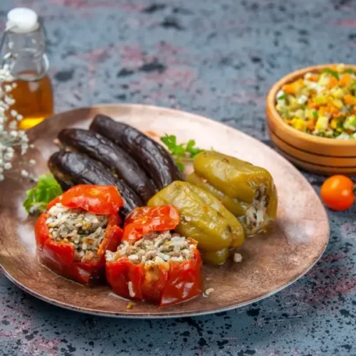 A platter of red and green stuffed peppers filled with rice and meat, served alongside stuffed eggplants and a bowl of chopped vegetable salad.