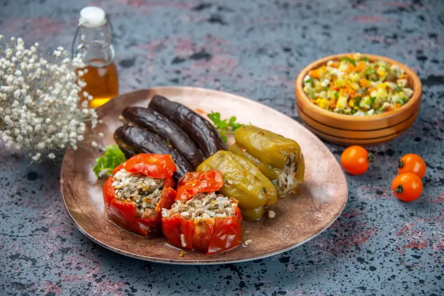 A platter of red and green stuffed peppers filled with rice and meat, served alongside stuffed eggplants and a bowl of chopped vegetable salad.