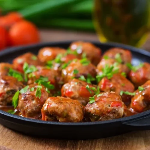 A cast-iron skillet filled with traditional Italian meatballs in rich tomato sauce, garnished with fresh parsley, with tomatoes and olive oil in the background.