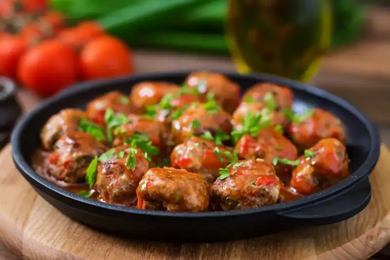 A cast-iron skillet filled with traditional Italian meatballs in rich tomato sauce, garnished with fresh parsley, with tomatoes and olive oil in the background.