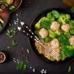 Bowl of quinoa with chicken meatballs, steamed broccoli, and fresh spinach, garnished with green onions and served on a dark background.