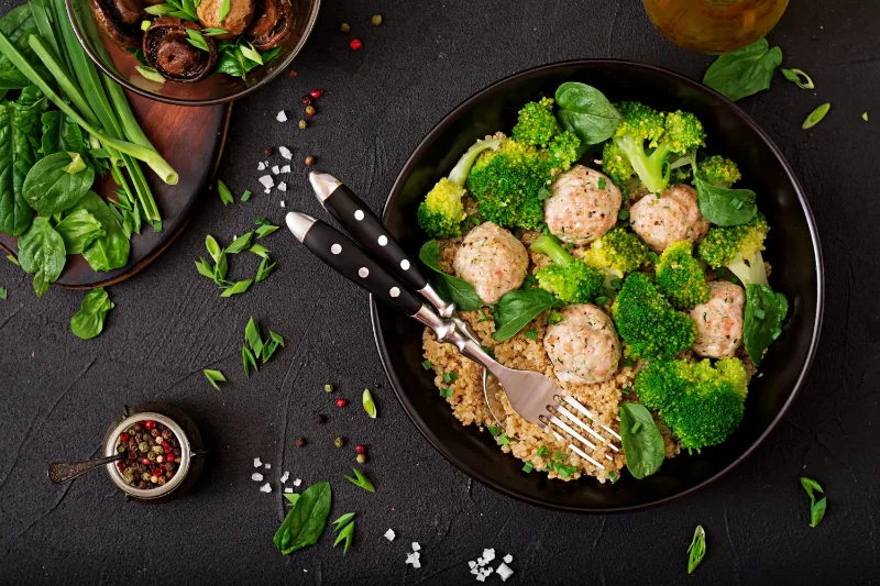 Bowl of quinoa with chicken meatballs, steamed broccoli, and fresh spinach, garnished with green onions and served on a dark background.