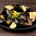 Steamed black mussels served on a dark plate with lemon wedges and chopped parsley on a wooden background.