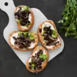 Four slices of mushroom bruschetta with a creamy spread, lettuce, and olives, served on a white cutting board with fresh parsley on a dark background.