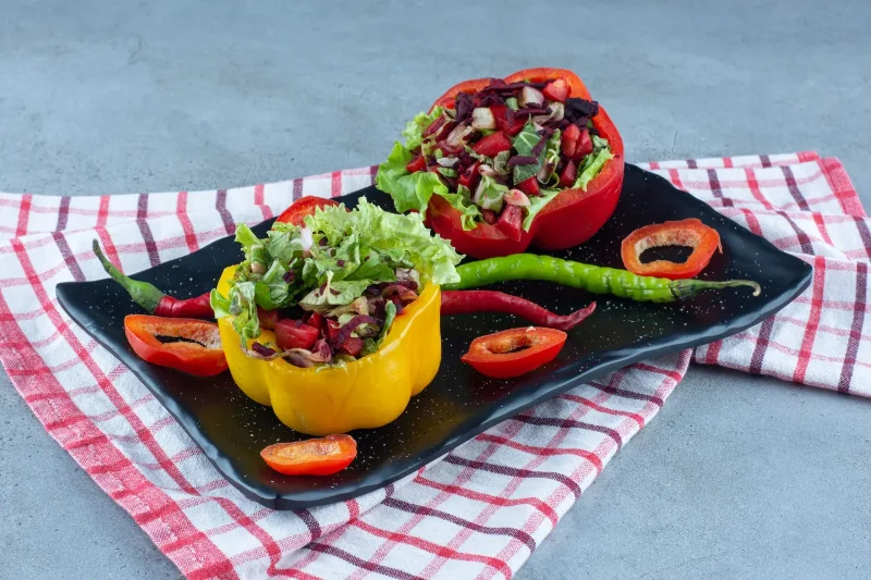 Red and yellow bell peppers stuffed with fresh lettuce, tomatoes, and chopped olives, garnished with green chili peppers and served on a black tray with a checkered cloth.