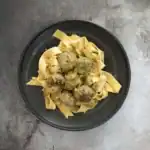 A plate of fettuccine pasta with meatballs in a creamy sauce, garnished with fresh herbs, served on a black plate.