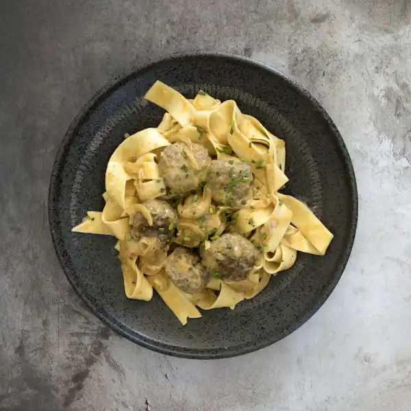 A plate of fettuccine pasta with meatballs in a creamy sauce, garnished with fresh herbs, served on a black plate.