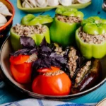 A pan filled with green bell peppers, tomatoes, and eggplants stuffed with a hearty vegetarian mixture, garnished with fresh basil, served alongside dumplings on a blue wooden table.