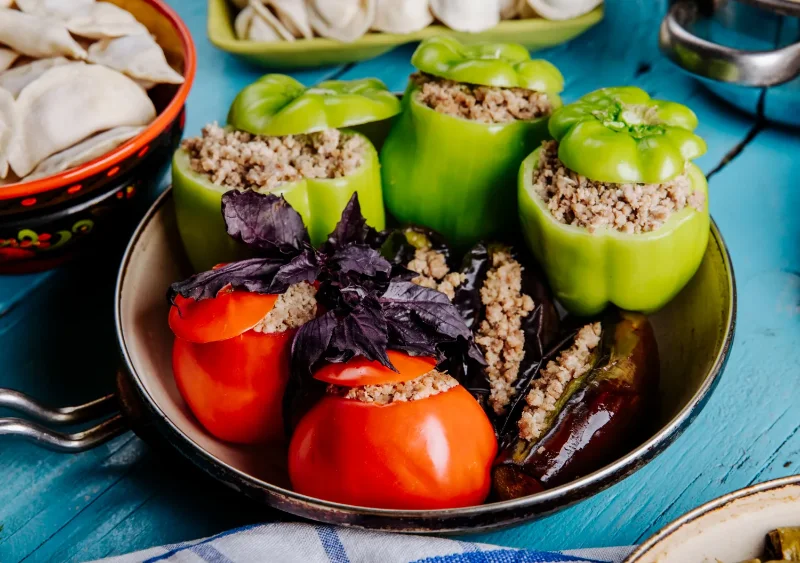 A pan filled with green bell peppers, tomatoes, and eggplants stuffed with a hearty vegetarian mixture, garnished with fresh basil, served alongside dumplings on a blue wooden table.