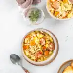 bowl of homemade sausage minestrone soup with sliced sausage, pasta, carrots, potatoes, and fresh herbs, served on a marble countertop with a spoon and a cozy napkin.