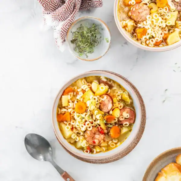 bowl of homemade sausage minestrone soup with sliced sausage, pasta, carrots, potatoes, and fresh herbs, served on a marble countertop with a spoon and a cozy napkin.