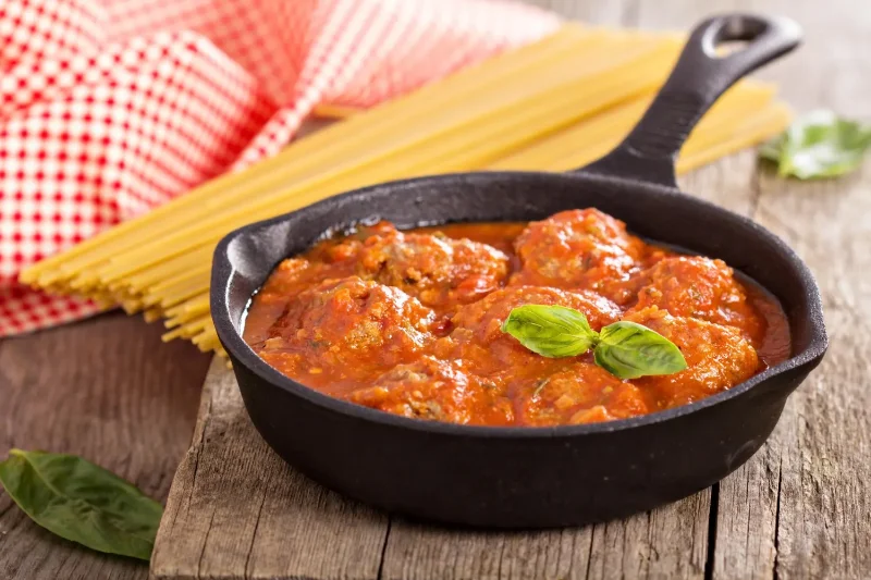 A cast-iron skillet filled with Italian meatballs in rich tomato sauce, garnished with fresh basil leaves. Uncooked spaghetti and basil leaves are placed nearby on a rustic wooden table.