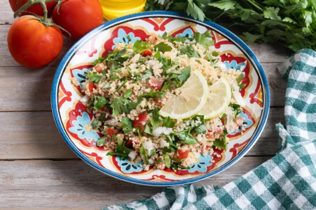 A colorful quinoa salad with fresh parsley, tomatoes, onions, and lemon slices in a vibrant patterned bowl, placed on a rustic wooden table.