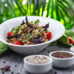 A fresh quinoa salad with cherry tomatoes, herbs, and greens in a white bowl, surrounded by spices and greenery.