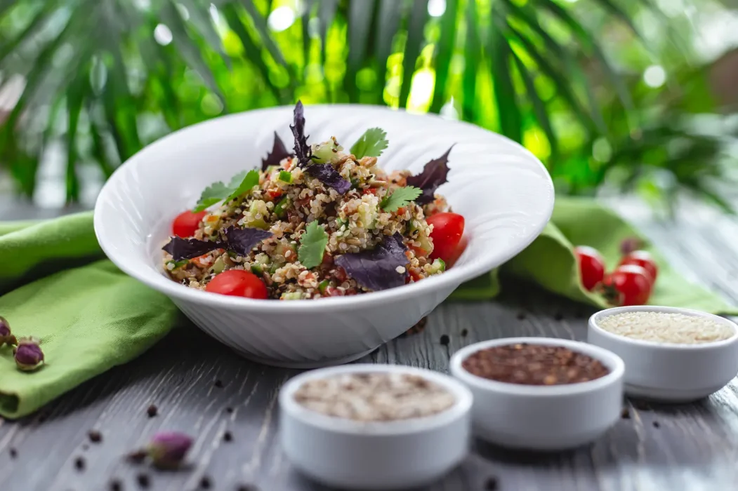 A fresh quinoa salad with cherry tomatoes, herbs, and greens in a white bowl, surrounded by spices and greenery.