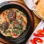 A black dish with baked meatball Shukshuka, featuring meatballs, tomatoes, herbs, and creamy sauce, served alongside sliced bread, cucumbers, and tomatoes.