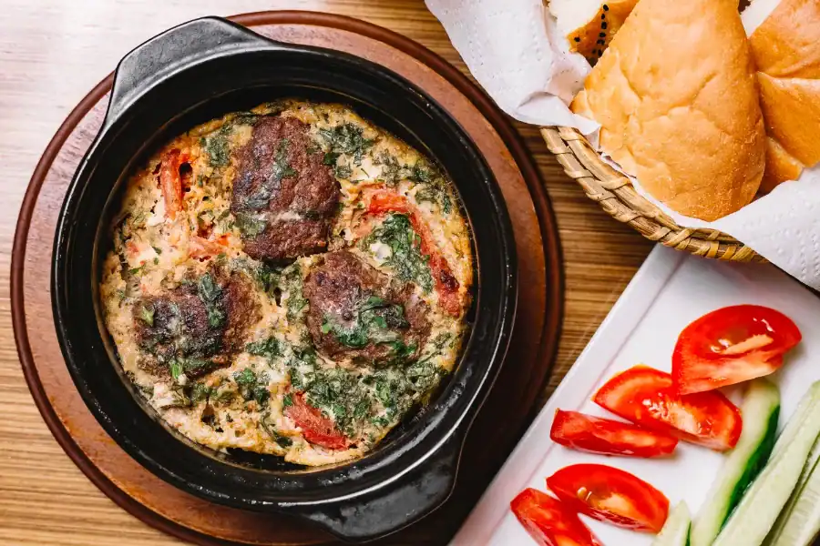 A black dish with baked meatball Shukshuka, featuring meatballs, tomatoes, herbs, and creamy sauce, served alongside sliced bread, cucumbers, and tomatoes.