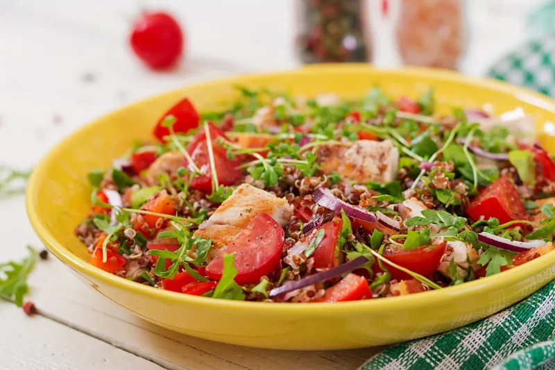 Colorful quinoa salad with grilled chicken, fresh tomatoes, red onions, and arugula, served in a vibrant yellow bowl.