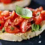 A close-up of a toasted bread slice topped with diced cherry tomatoes, fresh basil leaves, and olive oil, served on a dark surface.