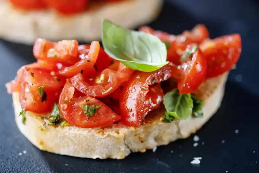 A close-up of a toasted bread slice topped with diced cherry tomatoes, fresh basil leaves, and olive oil, served on a dark surface.