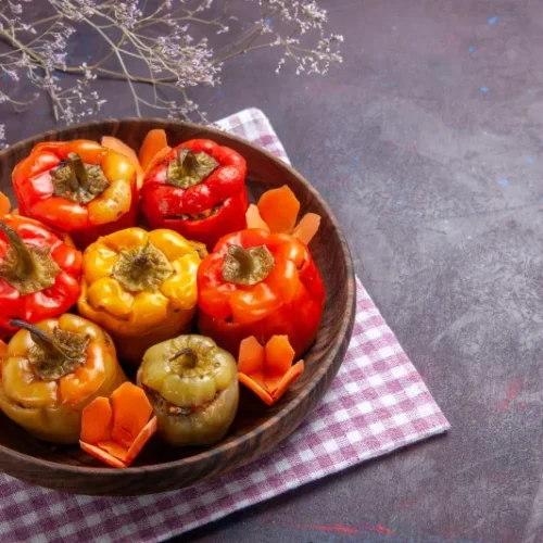 A wooden bowl filled with red, yellow, and green stuffed bell peppers, garnished with sliced carrots, set on a checkered cloth with dried flowers in the background.