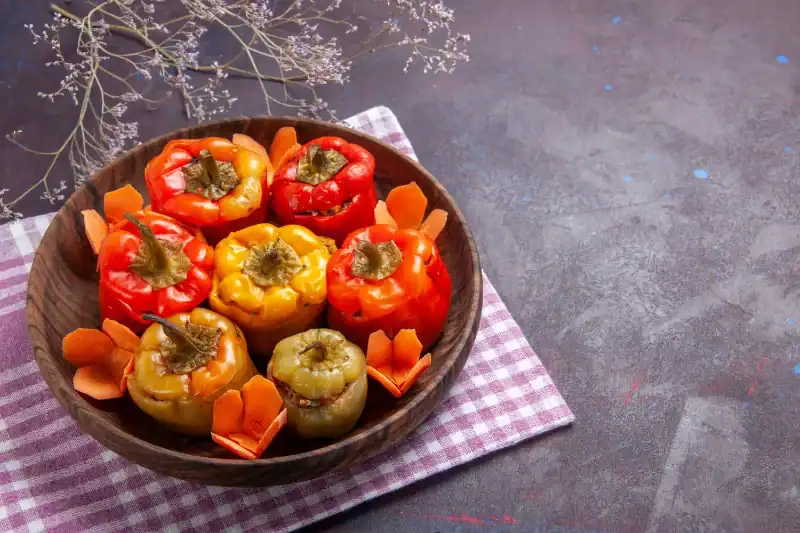 A wooden bowl filled with red, yellow, and green stuffed bell peppers, garnished with sliced carrots, set on a checkered cloth with dried flowers in the background.