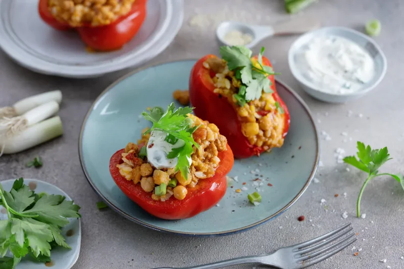 Red bell peppers stuffed with a mix of chickpeas, rice, and spices, garnished with fresh cilantro and a dollop of yogurt, served on a blue plate with additional herbs and sauce on the side.