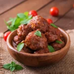 A wooden bowl filled with traditional Italian meatballs in rich tomato sauce, garnished with fresh basil leaves. Cherry tomatoes and basil sprigs are scattered on a rustic wooden table.