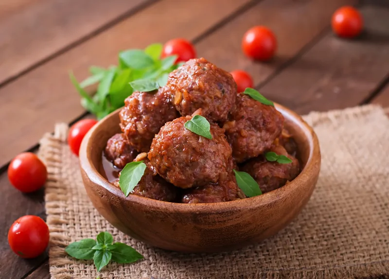 A wooden bowl filled with traditional Italian meatballs in rich tomato sauce, garnished with fresh basil leaves. Cherry tomatoes and basil sprigs are scattered on a rustic wooden table.