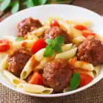 A close-up of a white plate filled with penne pasta, juicy meatballs covered in marinara sauce, cherry tomatoes, and fresh basil leaves.