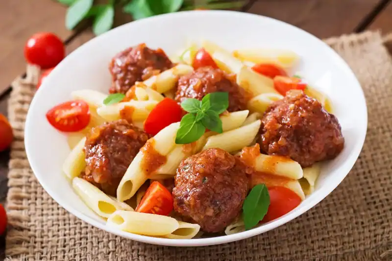 A close-up of a white plate filled with penne pasta, juicy meatballs covered in marinara sauce, cherry tomatoes, and fresh basil leaves.
