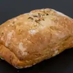 A freshly baked ciabatta bread with a golden crust, topped with sesame seeds, oats, and grains, placed on a black background.
